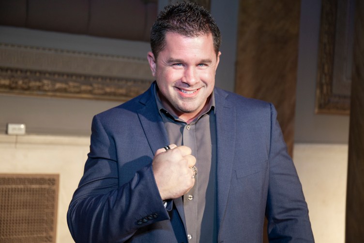 A man with gelled, spiky hair wears a suit and grins at the camera with a fist clenched in front of his chest.