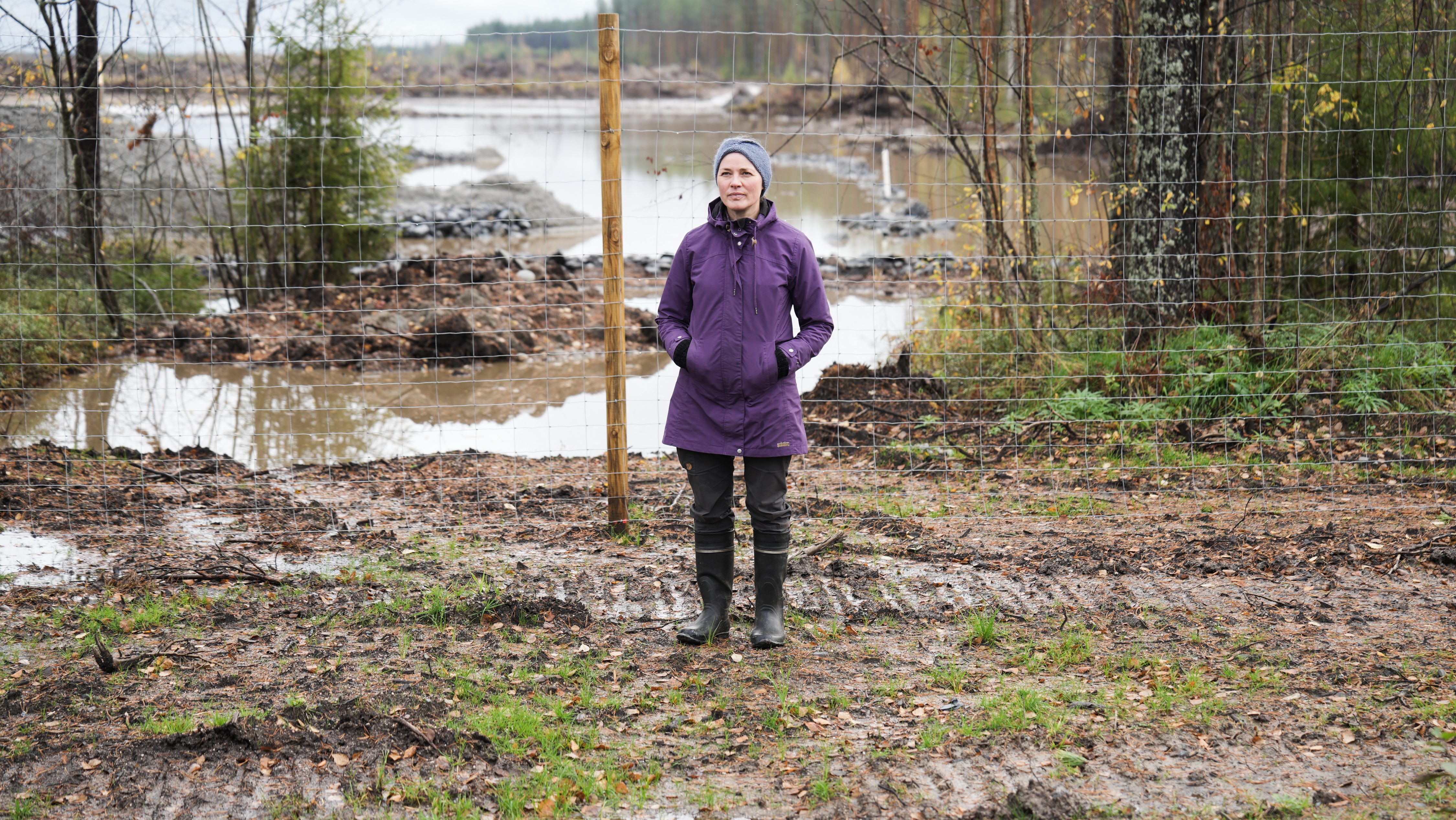 A woman in a purple coat stands in a muddy area in front of a wire fence.